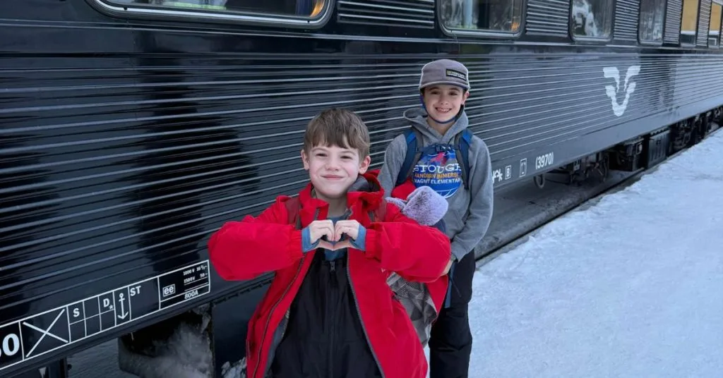 Two boys with backpacks on a snowy Narvik platform beside the SJ night train to Stockholm