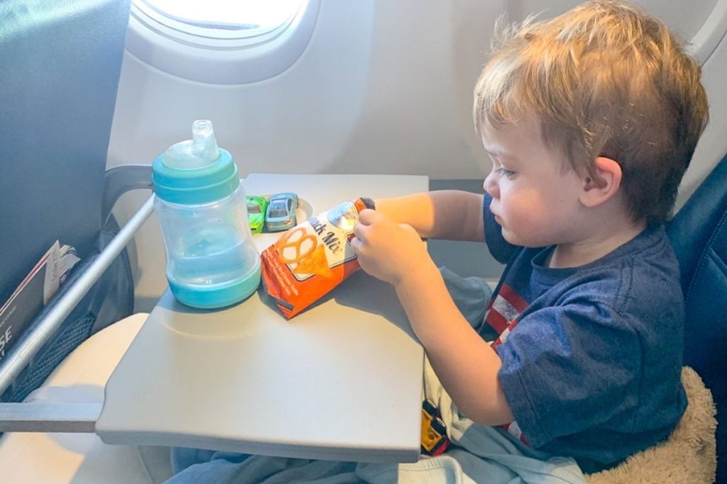 Toddler with sippy cup, snack pouches, and toy cars on airplane tray table during flight